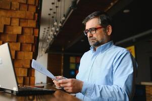 Positive senior bearded man with grey hair drinking coffee and using laptop at cafe, copy space. Stylish aged businessman in burgundy jacket enjoying his tea while working online. photo