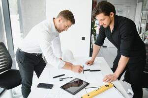 two building designers standing in a modern office leaning over a desk discussing blueprints together photo