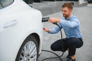 Hansome bearded guy sitting near his new modern electric car and holding plug of the charger, while car is charging at the charging station. photo