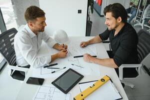 two building designers standing in a modern office leaning over a desk discussing blueprints together photo