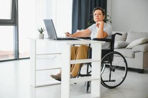 medio años mujer utilizando ordenador portátil sentado en silla de ruedas a hogar foto