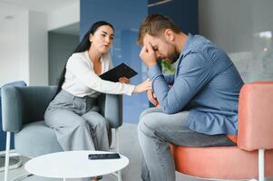 Handsome depressed man in casual clothes is leaning on his knees and telling about his problems while sitting on the couch at the psychotherapist. photo