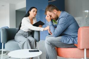 hermoso Deprimido hombre en casual ropa es propensión en su rodillas y narración acerca de su problemas mientras sentado en el sofá a el psicoterapeuta foto