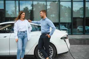 Happy young adult man and smiling woman charging electric car. photo