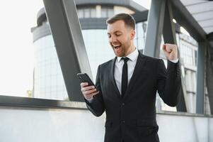 retrato de un emocionado joven empresario vestido en traje participación móvil teléfono, celebrando. foto