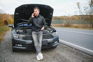 hermoso joven hombre vocación para asistencia con su coche roto abajo por el borde del camino foto