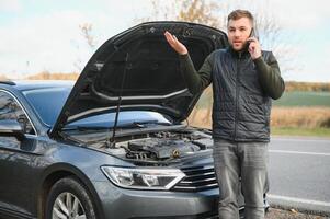 Man repairing a broken car by the road. Man having trouble with his broken car on the highway roadside. Man looking under the car hood. Car breaks down on the autobahn. Roadside assistance concept. photo
