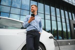 Man charging his electric car at charge station and using smartphone photo