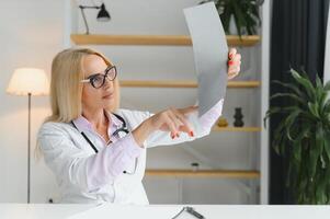 retrato de medio años hembra médico es vistiendo un blanco del doctor Saco con un estetoscopio alrededor su cuello. sonriente médico en pie a privado clínica foto