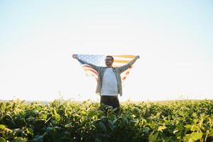 un joven granjero soportes con un Estados Unidos bandera en un haba de soja campo. el concepto de el nosotros agrícola industria. foto