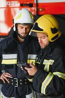 Two professional firefighters with uniforms and protective helmets posing infront of a firetruck. photo