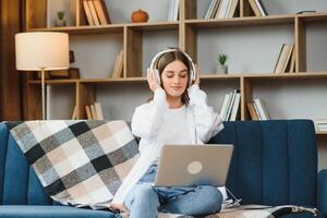 Beautiful young caucasian woman in casual clothing enjoying music and smiling while resting at home. Young woman with headphones using laptop at home photo