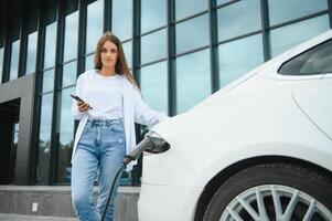 Phone in hands. Woman on the electric cars charge station at daytime. Brand new vehicle. photo