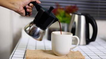 A woman hand pours black coffee from a moka pot into a white mug, kitchen interior. Front view, selective focus. video