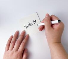 Positive vibes message saying Smile handwriting on top of square paper with hands holding the paper and black marker. Object photography isolated on white studio background. photo