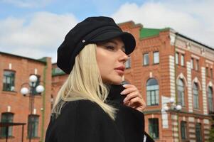 hermosa de moda joven rubia caucásico mujer en un elegante negro gorra y Saco en contra el antecedentes de ladrillo ciudad edificios foto