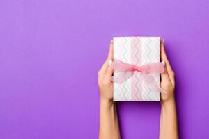 Flat lay of woman hands holding gift wrapped and decorated with bow on purple background with copy space. Christmas and holiday concept photo