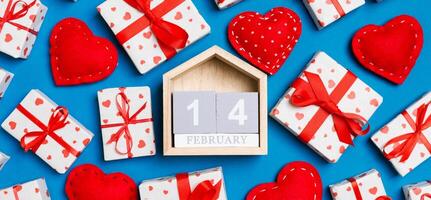 Composition of wooden calendar, holiday white gift boxes and red textile hearts on colorful background. The fourteenth of February. Valentine's day concept photo