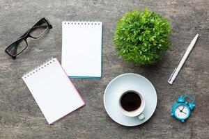 moderno oficina escritorio mesa con cuaderno y otro suministros con taza de café. blanco cuaderno página para usted diseño. parte superior vista, plano laico foto