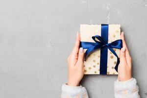 Flat lay of woman hands holding gift wrapped and decorated with bow on gray cement wooden background with copy space. Christmas and holiday concept photo