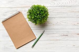 moderno oficina escritorio mesa con cuaderno y otro suministros con verde árbol. blanco cuaderno página para usted diseño. parte superior vista, plano laico foto