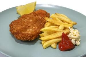 Breaded steak with French fries, with ketchup and mayonnaise, two lemon wedges, served on a turquoise plate. Isolated in a white background. photo