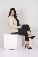 a woman sitting on a white cube wearing white jacket and black heels photo