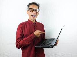 retrato de un asiático hombre vistiendo un rojo ropa participación un ordenador portátil con un sonriente cara expresión. foto