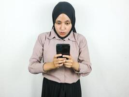 surprised expression of a young Asian woman wearing a hijab when looking at a smart phone screen photo