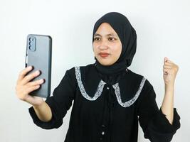 emocionado hermosa asiático mujer en hijab utilizando un móvil teléfono apretando puño recibido bueno Noticias foto