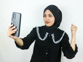 Excited beautiful Asian woman in hijab using a mobile phone clenching fist received good news photo