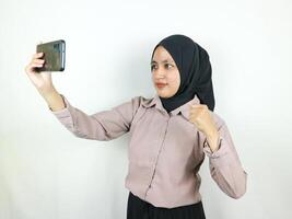 emocionado hermosa asiático mujer en hijab utilizando un móvil teléfono apretando puño recibido bueno Noticias foto