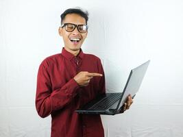 retrato de un asiático hombre vistiendo un rojo ropa participación un ordenador portátil con un sonriente cara expresión. foto