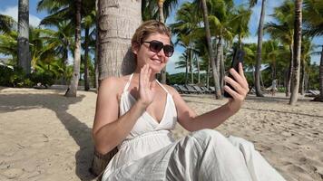 contento mujer en Gafas de sol utilizando teléfono inteligente en tropical playa con palma árboles, simbolizando ocio viaje y verano vacaciones video