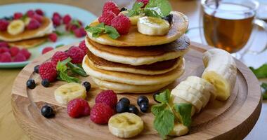 Sweet honey pouring over pancakes. Tasty breakfast food. Pancakes are served with raspberries, banana, blueberry and mint leaf. Honey dripping down on dessert. Closeup Ultra 4K video