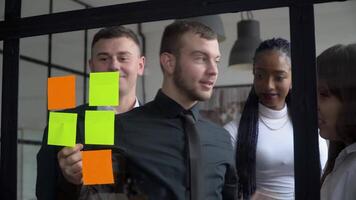 Corporate business multiethnic people using sticky notes, solving strategy on glass whiteboard. Team leader male showing solution for project deadline in office meeting video