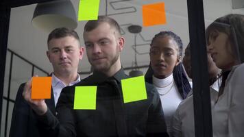 Corporate business multiethnic people using sticky notes, solving strategy on glass whiteboard. Team leader male showing solution for project deadline in office meeting video