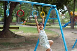 contento niña colgando en mono bar por mano haciendo ejercicio. pequeño asiático niña jugando a al aire libre patio de recreo en el parque en verano vacaciones. sano actividad. foto