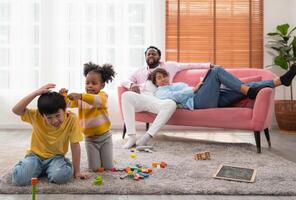 niños jugar con de madera juguete en piso en vivo habitación mientras padre sentado en sofá. dos niños jugando con vistoso bloques foto