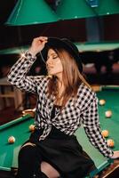 un niña en un sombrero en un de billar club se sienta en un de billar mesa.jugando piscina foto