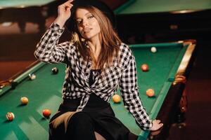 A girl in a hat in a billiard club sits on a billiard table.Playing pool photo