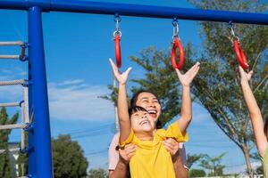 Cute little girl hanging bars in the playground. Happy little Asian child having fun on playground, climbing bars in park. Little girl playing in outdoor playground with mother. photo