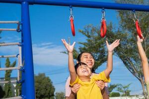 Cute little girl hanging bars in the playground. Happy little Asian child having fun on playground, climbing bars in park. Little girl playing in outdoor playground with mother. photo