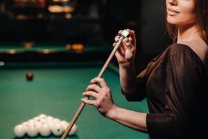 A girl in a dress stands with a cue in her hands and cleans it with chalk in a billiard club with balls in her hands.Playing billiards photo