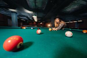 A girl in a hat in a billiard club with a cue in her hands hits a ball.Playing pool photo