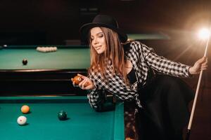 A girl in a hat in a billiard club with a cue and balls in her hands.Playing pool photo