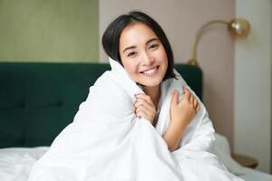 Cute and tender woman sitting in bed with warm duvet, smiling as looking out of the window, enjoying morning and comfort of blanket photo