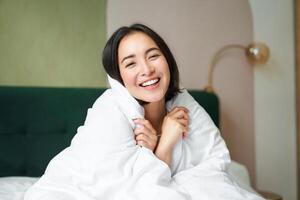 Beautiful romantic woman covers herself with warm duvet, enjoys sleeping and staying in bed, waking up happy in morning, sitting in bedroom photo