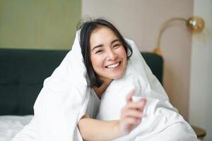 hermosa asiático mujer sentado en cama, cubierto con blanco funda Nordica, sonriente, disfrutando contento fin de semana mañana, riendo a cámara foto