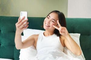 Positive girl blogger takes selfie in her bed, bedroom photo on smartphone, smiling and showing peace v-sign gesture